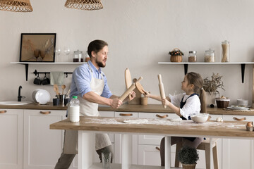 Happy young daddy and little adorable daughter wear aprons holding wooden appliances fighting,...