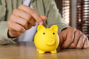 Man putting coin into yellow piggy bank on wooden table, closeup