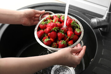 Woman washing fresh strawberries under tap water in metal colander above sink, closeup