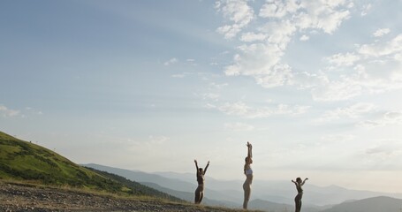 Three young women perform synchronized yoga poses on a mountain ridge, their movements harmonized...