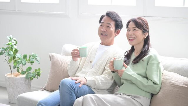 リビングでテレビを見ながら飲み物やコーヒーを飲む50代の夫婦　会話をするなどポシティブなイメージの動画　長袖