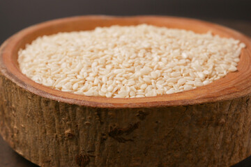 A wooden bowl filled with white sesame seeds, accompanied by a spoon, set on a dark surface