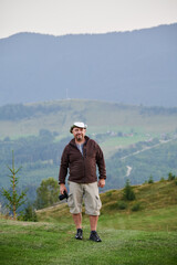 Male tourist hiking in the mountains, traveling in summer. Man standing on hill, holding camera,...