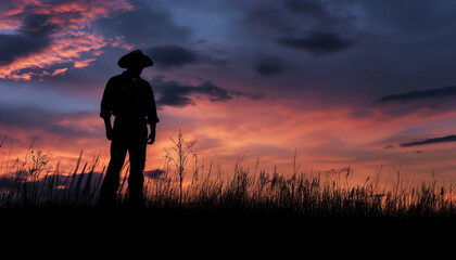 Fototapeta premium Silhouette of a man wearing a hat standing in a field at sunset with a dramatic sky