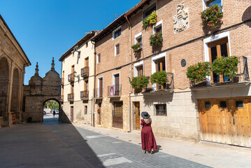 Blonde woman with long hair, red dress and hat strolling through the streets of the medieval tourist village of Los Arcos in Navarre