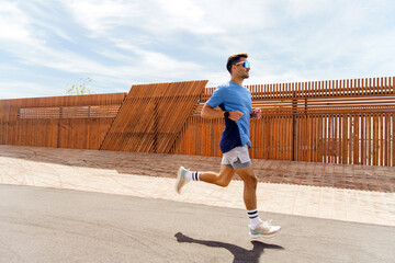 Man in a blue shirt running along a modern wooden fence, maintaining a brisk pace under a clear sky.