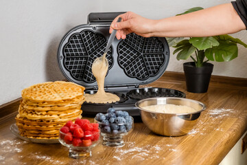 Making Belgian waffles. Female hand pours dough into waffle maker in kitchen at home. Cooking summer breakfast with round waffles and berries