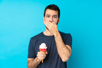 Young man with a cornet ice cream over isolated blue background covering mouth with hands