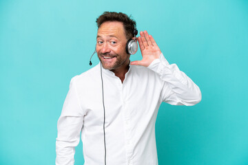 Telemarketer caucasian man working with a headset isolated on blue background listening to something by putting hand on the ear