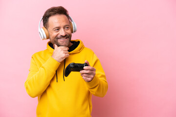 Middle age man playing with a video game controller isolated on pink background looking to the side and smiling