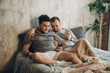 Obraz premium Handsome homosexual couple relaxing in bed before night sleep and looking at smart phone screen, making video conference call, talking to their friends online. People, relationships and technology.
