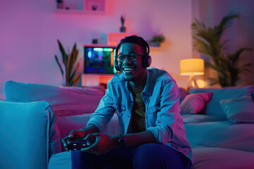 Young African American gamer with gamepad wearing headphones plays a console game in a cozy, neon-lit room, enjoying a relaxed gaming session.