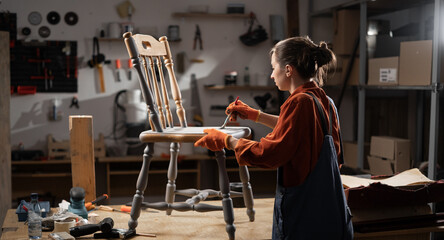 Young woman painting old wooden chair in grey color with paint brush working in her workshop....