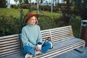 Smiling blonde stylish mature woman 50-55 year old wearing hat and knitted sweater over sitting on bench and drinking coffee outdoor in park. Autumn season.