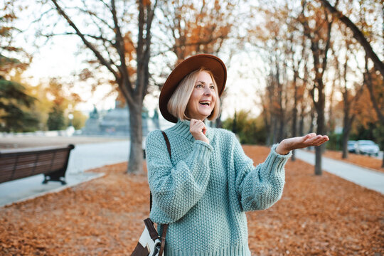 Smiling blonde stylish mature woman 50-55 year old wearing hat and knitted sweater over fallen leaves in park outdoor. Happiness. Autumn season.