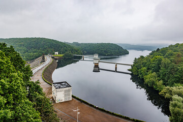 Fototapeta premium Aerial view of Gileppe Dam, wall with pedestrian path on crest, two intake or outlet towers protruding from lake waters, lush trees in valley mountains covered in fog, cloudy day in Jalhay, Belgium