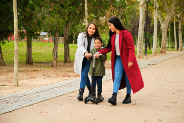 Portrait of Latin lesbian couple helping their son to ride a scooter in a park. LGBT family.