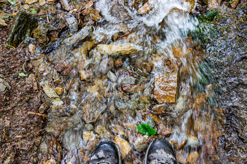 Low level water flowing quickly over stones, toes cap of two male hiking boots in river water, man standing on shore on a summer day
