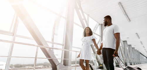 Fototapeta premium A happy couple walks hand-in-hand through a bright airport terminal, pulling luggage behind them. The man is wearing a white t-shirt and jeans, while the woman is wearing a white dress.