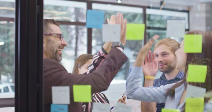 Work colleagues discussing startup project ideas and giving high five. Caucasian business team at glass wall with sticky notes. Scrum with colorful stickers. Vertical movement.