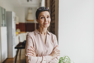 Charming pretty stylish real estate agent or home owner in elegant shirt with glasses on head smiling at camera standing with crossed hands in front of window, waiting for prospective clients