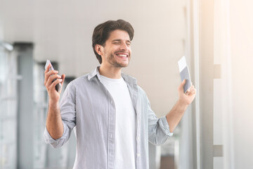 Enjoying Travels. Portrait Of Happy Young Man Standing In Airport, Holding Passport With Tickets And Smartphone In Hands, Raising Hands In Excitement