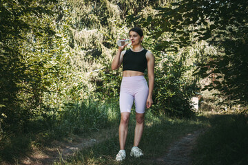 A young sporty woman drinking water from a bottle in the summer park, morning workout and hydration concept