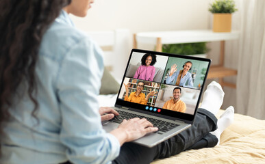 A woman sits on a bed in her home, using a laptop to video chat with friends. She is smiling and typing on the keyboard. The laptop screen shows four people in different video windows
