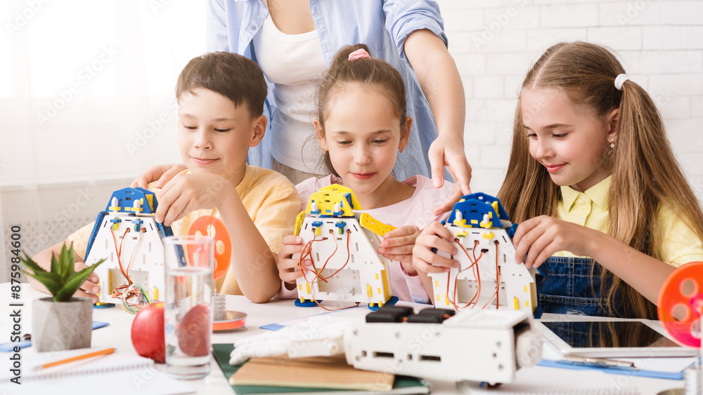 Poster A group of young students are working together to build robots in a classroom setting. They are being guided by a teacher, who is standing behind them and observing their progress.
