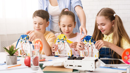 A group of young students are working together to build robots in a classroom setting. They are being guided by a teacher, who is standing behind them and observing their progress.