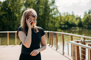 A happy blonde woman talking on a mobile phone by the river. Technology, communication and people concept. Positive 30s female speak phone on nature background,