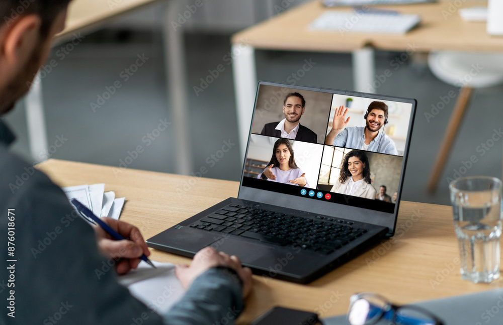 Poster A man sits at a desk in an office setting, taking notes on a notepad while attending a video conference meeting with four colleagues. The conference is displayed on a laptop screen