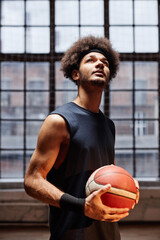 Vertical portrait of young African American man as a basketball player holding ball and looking up in dramatic lighting against window