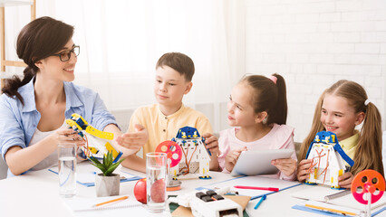 A teacher is showing three students how to build a model robot in a classroom setting. The students are all engaged and interested in the activity. The teacher is pointing to a part of the robot