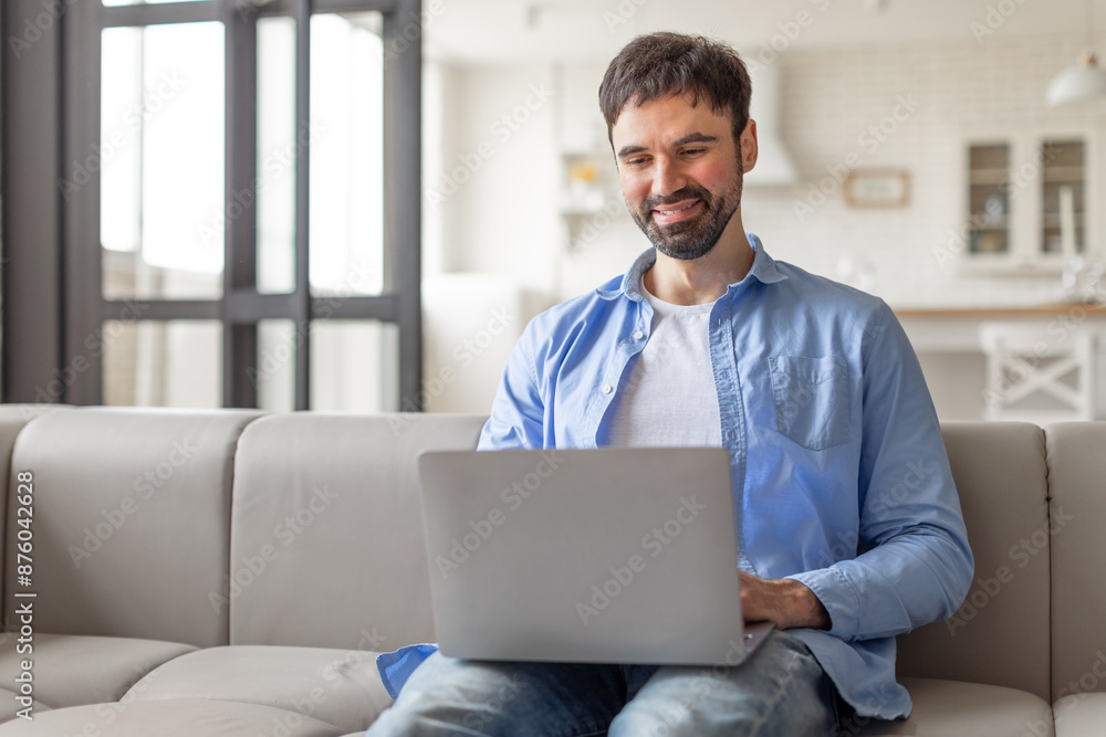 Poster a man sits on a grey couch in a modern living room and uses a laptop. he is smiling as he looks at t