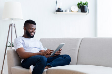 Black man is sitting on a light gray sofa in a living room, looking at a tablet. He is smiling and wearing a white t-shirt and blue jeans. There is a floor lamp and a plant in the background.