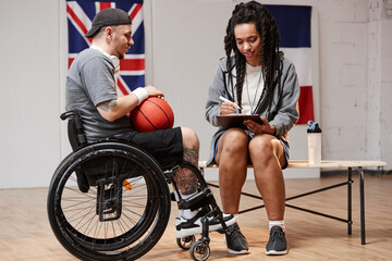 Side view portrait of smiling young sportsman using wheelchair and giving interview to girl with clipboard at indoor basketball court copy space