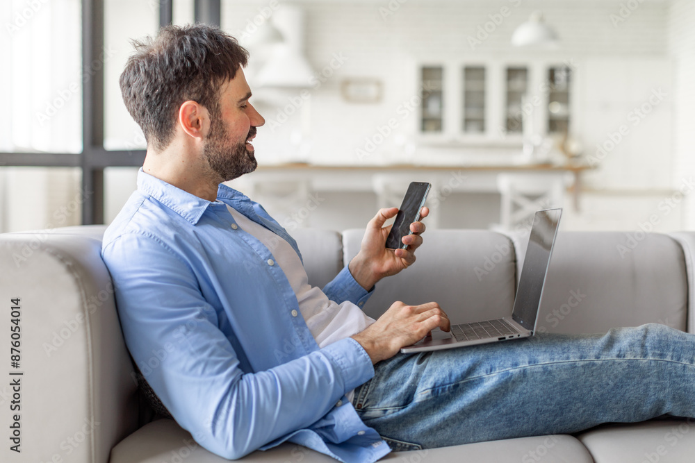 Poster A man sits on a couch in a modern home, comfortably using a smartphone and a laptop. He is smiling, likely engaging with a digital task or enjoying some entertainment.