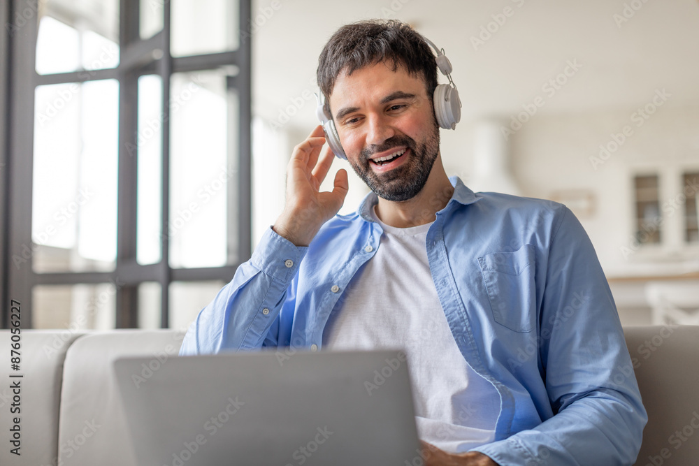 Poster A man is sitting on a couch with a laptop in front of him. He is smiling and appears to be video chatting while wearing headphones.