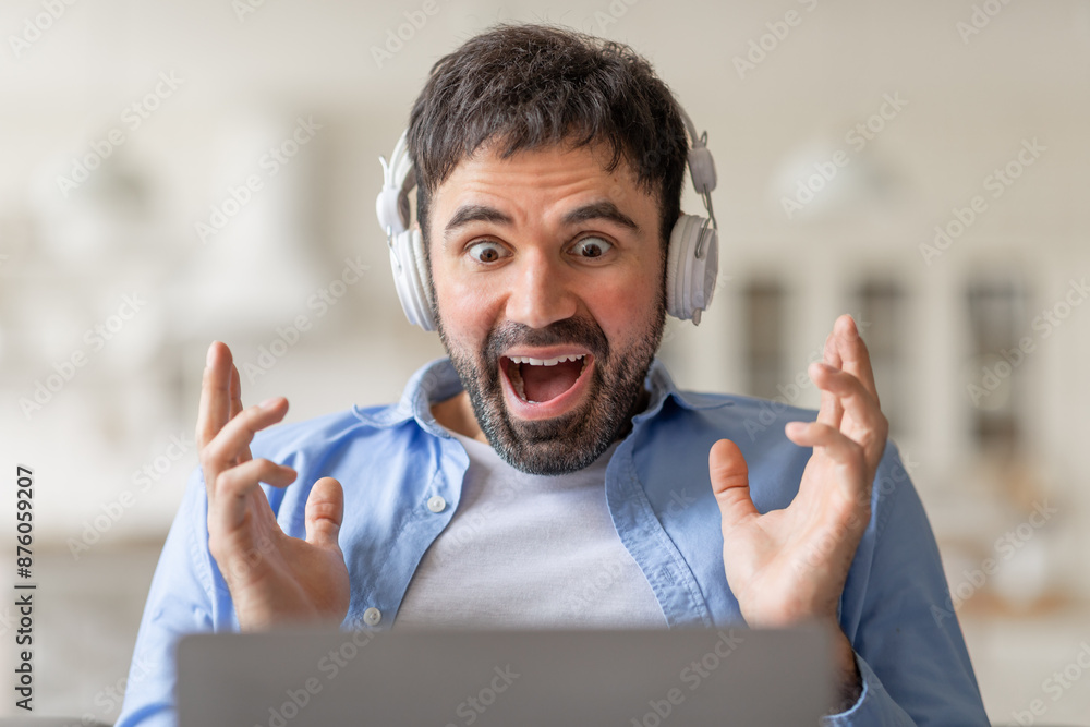 Poster a man wearing headphones sits in front of a laptop with a wide open mouth and his hands raised in th