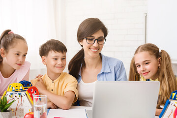 A teacher and three young students are gathered around a laptop computer in a classroom setting. They appear to be learning about robotics. The teacher is smiling and pointing at the screen