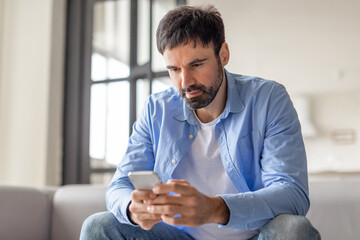 A man sits on a couch in his home, looking down at his smartphone. He is wearing a blue button-down shirt over a white t-shirt and jeans