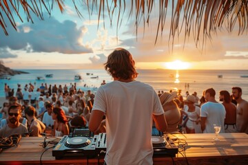 A DJ wearing headphones spins music at a beach party as the sun sets in the distance.