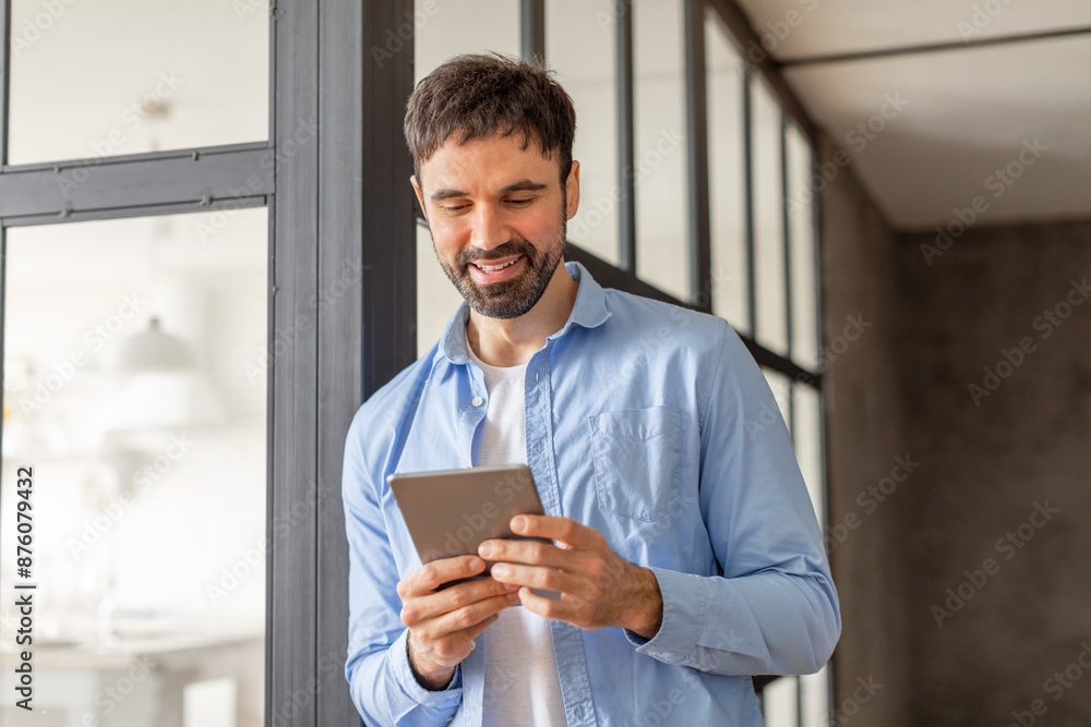 Poster A man in a blue shirt stands in an office building, using a tablet device while looking at the screen with a smile. The man is in a casual work environment, surrounded by large windows.