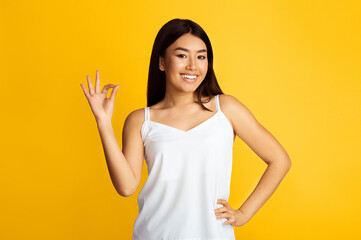 A young Asian woman with long black hair is smiling and making the okay sign with her right hand. She is wearing a white tank top and standing in front of a bright yellow background.