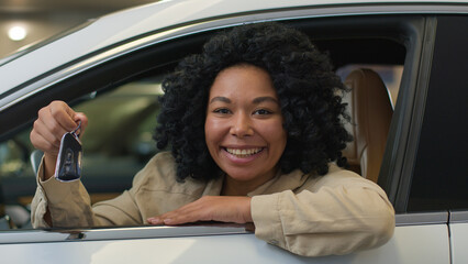 Happy African American girl female buyer client in new car showing key of luxury automobile...