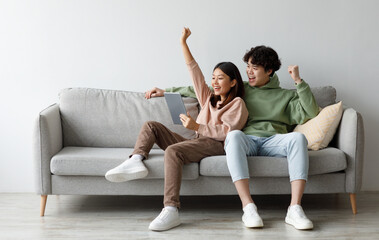 Excited young Asian couple sitting on couch with tablet pc, celebrating online win, great deal or...