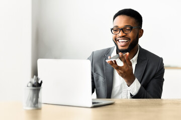 A businessman is sitting at his desk, using a voice assistant on his phone while looking at his laptop.