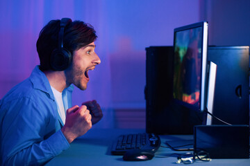 A young man wearing headphones sits at his computer, excitedly playing a video game. His mouth is open in surprise and his fists are clenched in victory as he watches the action unfold on the screen.