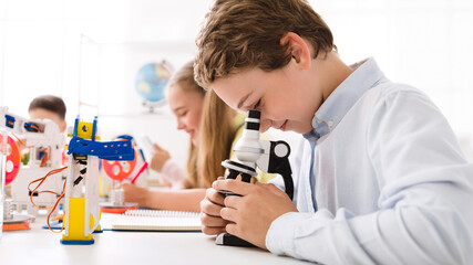 A young boy, wearing a white shirt, looks intently through a microscope, focusing on a slide. He is sitting at a desk in a classroom setting, surrounded by other students and educational materials